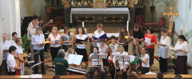Eine Gruppe von Menschen steht vor einem Kirchenchor, einige sitzen auf Bänken und andere stehen mit Büchern, während eine Person ein Instrument auf einem Stuhl spielt; der Hintergrund enthält Blumentöpfe, Kerzenständer, ein Kreuz und andere Gegenstände.