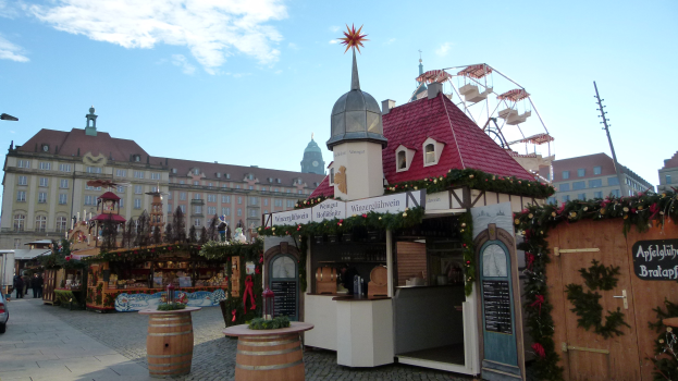 Ein belebter Weihnachtsmarkt auf einem Stadtplatz mit geschmückten Bäumen, Gebäuden, Straßenlaternen, Fahrzeugen, Passanten und einer bewölkten Himmel.