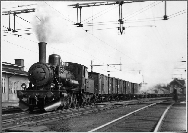 Schwarzes und weißes Foto eines Dampflokomotivs, das Rauch auf den Bahnschienen ausstößt, mit einem Gebäude links und Strommasten und -leitungen im Hintergrund.