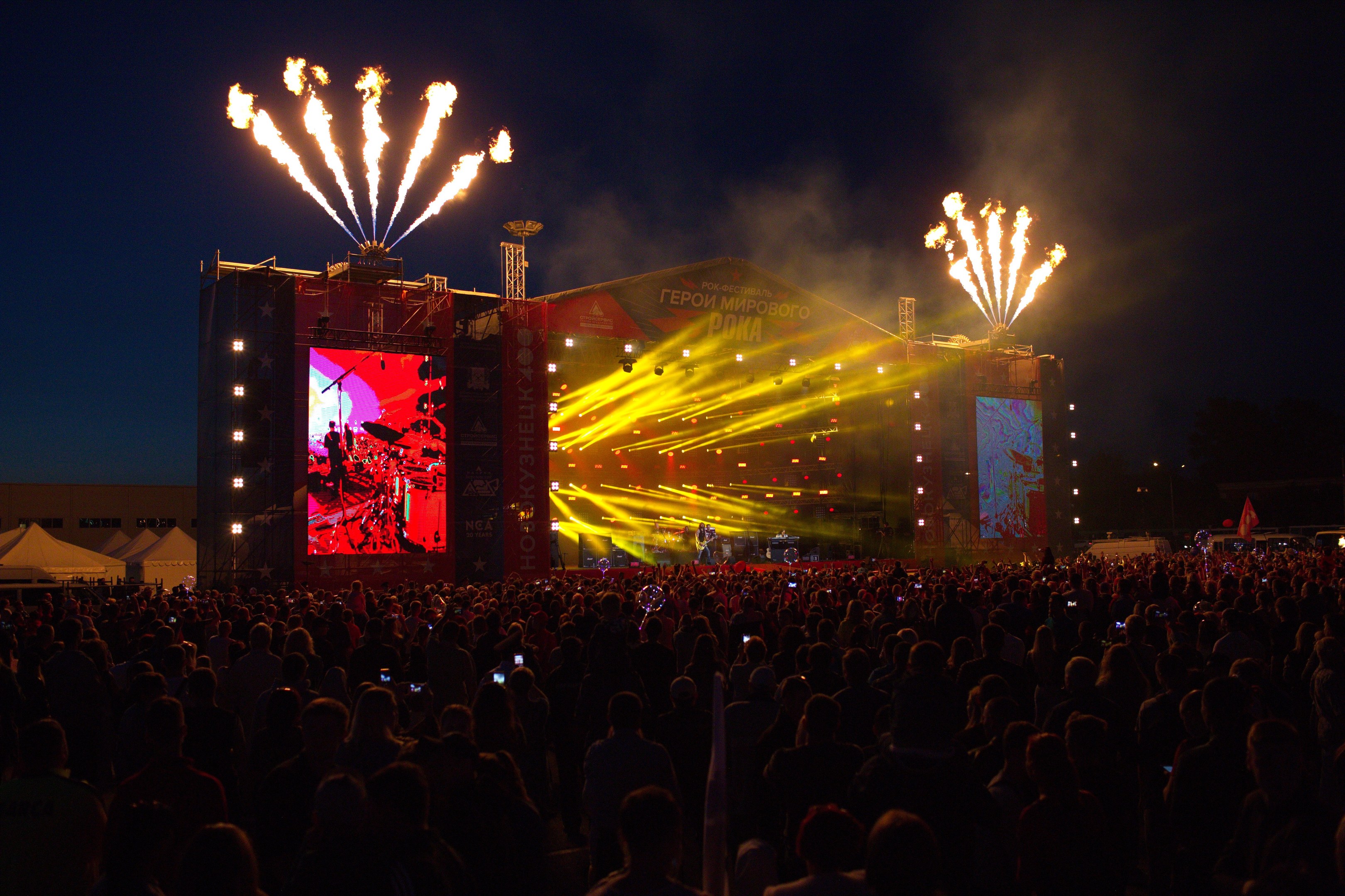 Eine große Menschenmenge bei einem Musikfestival mit beleuchteter Bühne, Performern, Bannern, Zelten links und Feuerwerk am dunklen Himmel.