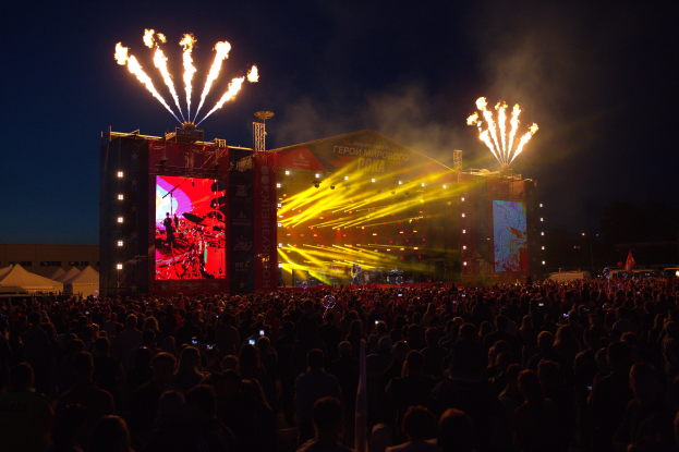 Eine große Menschenmenge bei einem Musikfestival mit beleuchteter Bühne, Performern, Bannern, Zelten links und Feuerwerk am dunklen Himmel.