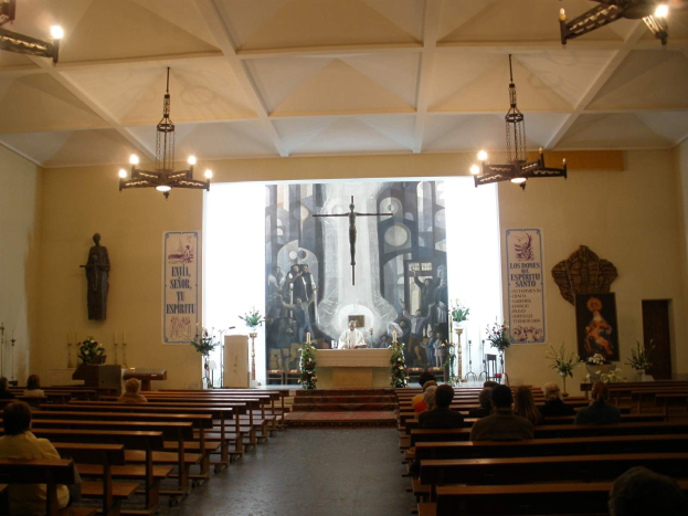 Eine Kircheninnenansicht mit Menschen in Kirchenbänken, die einem großen Bildschirm zugewandt sind, beleuchtet von Kronleuchtern, mit Blumenbouquets, Kerzenständern, Postern an den Wänden und einer Person in der Nähe eines Podiums mit einem Mikrofon.
