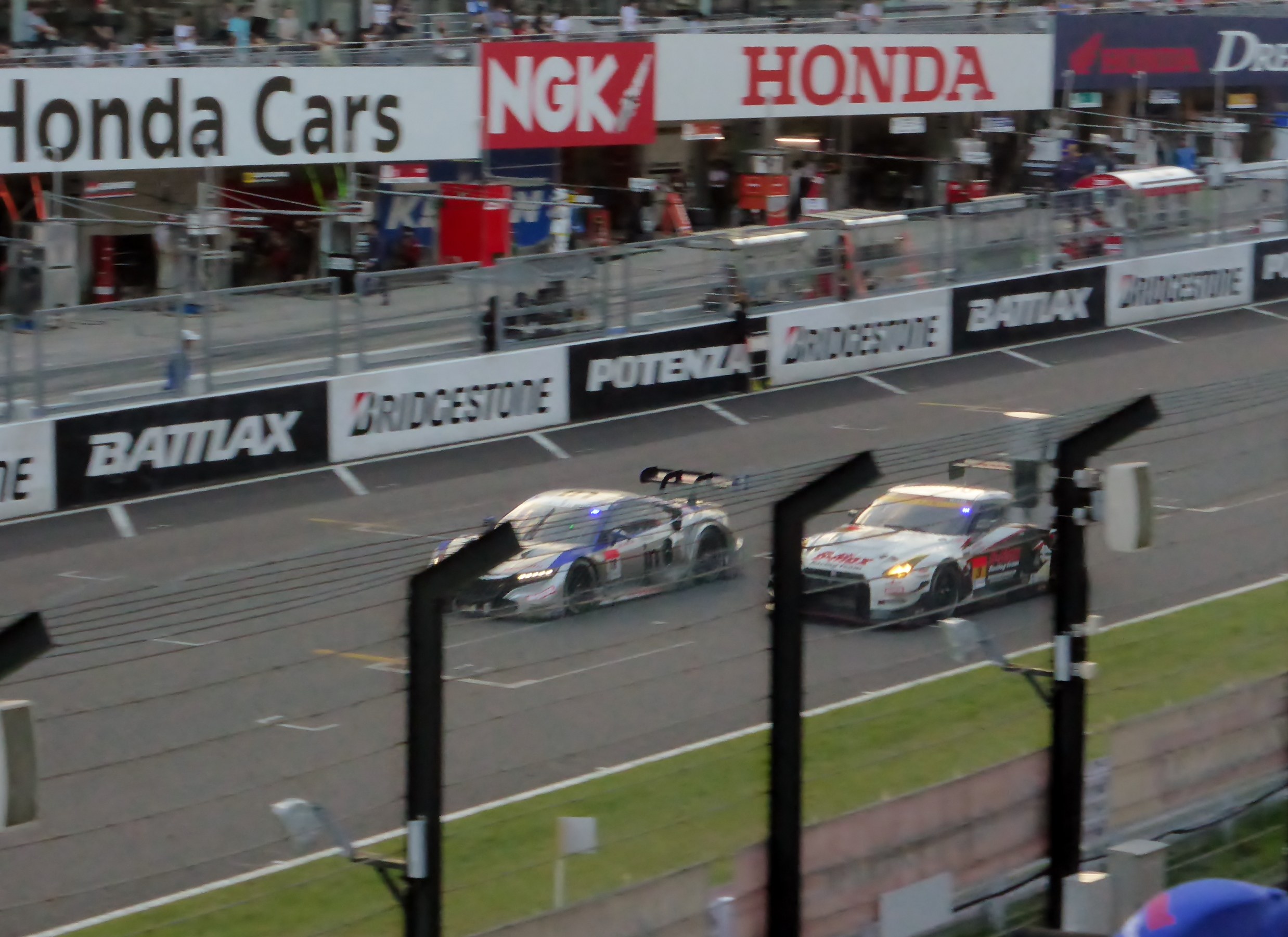 Gruppe von Autos auf einer Rennstrecke mit Zäunen, Schildern, Pfosten und einem Gebäude mit Fenstern.