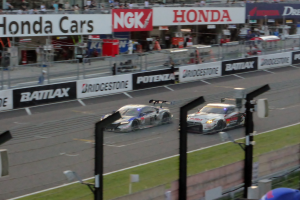 Gruppe von Autos auf einer Rennstrecke mit Zäunen, Schildern, Pfosten und einem Gebäude mit Fenstern.