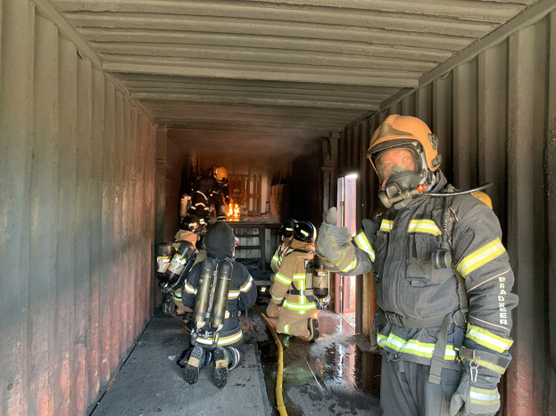 Feuerwehrleute in Schutzausrüstung in einem Seecontainer, einer h├Ąlt ein Rohr, mit einem Feuerl├Âscher im Hintergrund.