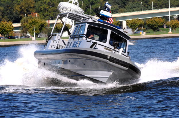 Polizeiboot fährt über ein Gewässer mit einer Person an Bord, Brücke und Bäume sind im Hintergrund unter einem klaren blauen Himmel zu sehen.