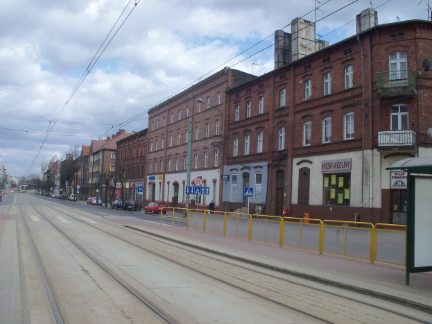 Stadtstraße mit einer Bahn auf Schienen, umgeben von Gebäuden, Schildern, Laternen, Strommasten mit Drähten, Bäumen und einem bewölkten Himmel.