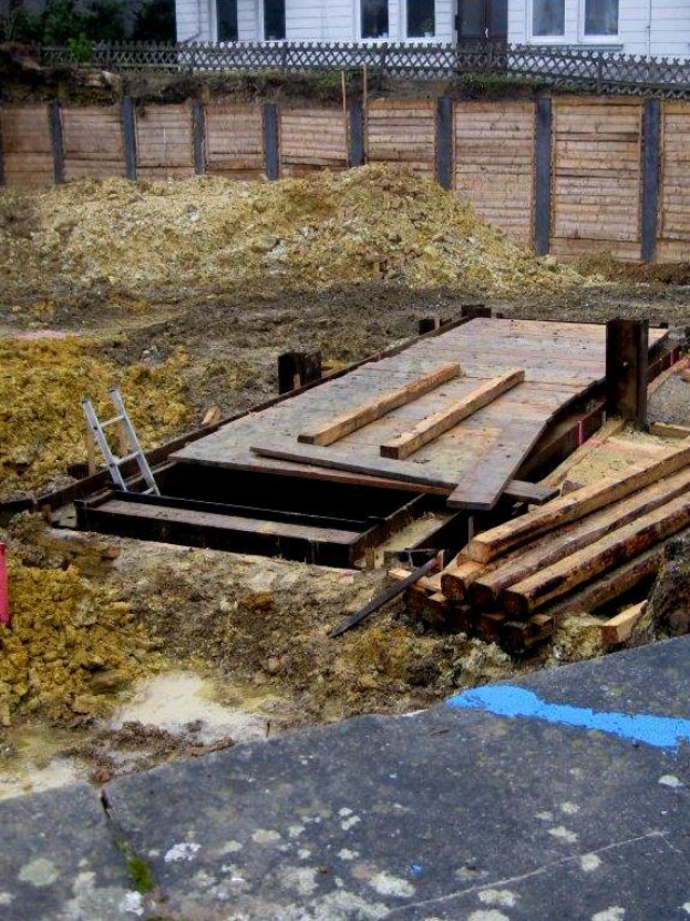 Haus im Bau mit hölzernen Brettern und einer Leiter im Vordergrund, umgeben von Bäumen und einem Zaun, mit einem Haufen Erde in der Nähe.