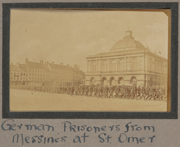 Deutsche Kriegsgefangene aus Messines vor einem Gebäude mit einem klaren blauen Himmel im Hintergrund, unten sichtbarer Text.