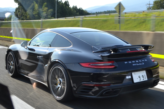Ein schwarzer Porsche 911 GTS fährt auf einer Autobahn, umgeben von einem Zaun, Gras, Bäumen, Pfählen, Drähten und Bergen unter einem klaren blauen Himmel.
