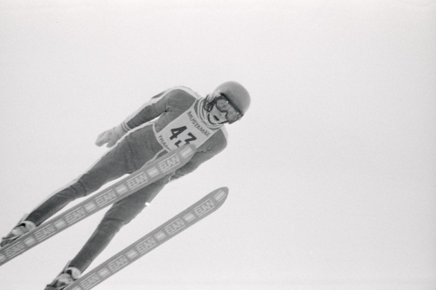 Skispringer in voller Ausr├╝stung springt in der Luft auf Skiern gegen einen klaren blauen Himmel