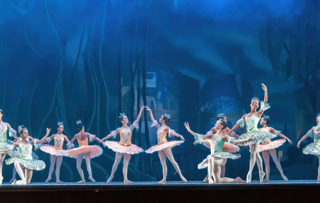 Eine Gruppe von Balletttänzerinnen in farbenfrohen Kostümen, die *Der Nussknacker* auf der Bühne des Royal Opera House tanzen, bewegen sich synchronisiert vor einem blauen Hintergrund.