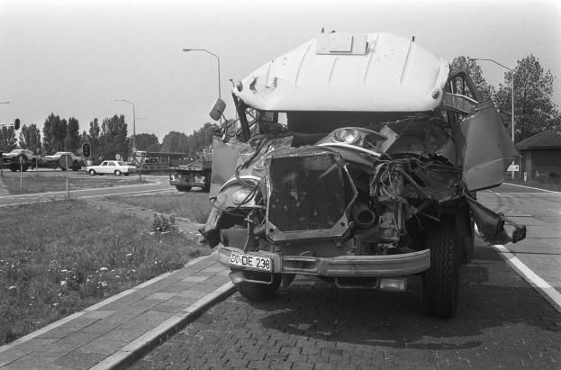 Schwarzes und weißes Foto eines stark beschädigten Autos am Straßenrand mit eingedellter Motorhaube und zersplitterten Fenstern, umgeben von anderen Fahrzeugen, Laternenpfählen, Bäumen und einem Haus.