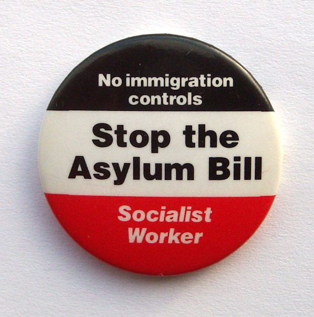 Ein rundes schwarzes Abzeichen mit weißer fettgeschriebener Schrift "Stoppt das Asylgesetz Sozialistische Arbeiterin" auf einer weißen Fläche.
