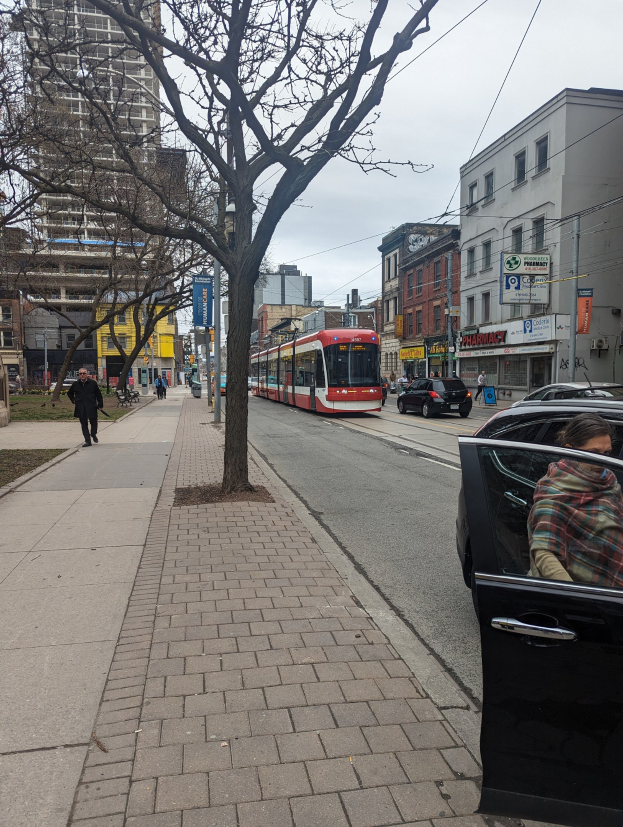 Eine Frau steigt aus einem Auto auf einer Stadtstraße mit einer Straßenbahn im Hintergrund, Bäume säumen die Straße, Gebäude mit Fenstern, Texttafeln, Fußgänger auf dem Gehweg und sichtbarer Himmel.
