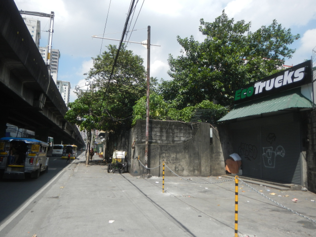 Stadtstraße mit Fahrzeugen, eine Brücke auf der linken Seite, Gebäude, Bäume, Strommasten, Drähte und ein bewölkter Himmel, mit einem Gebäude auf der rechten Seite mit einer 'Eco-Trucks'-Aufschrift.
