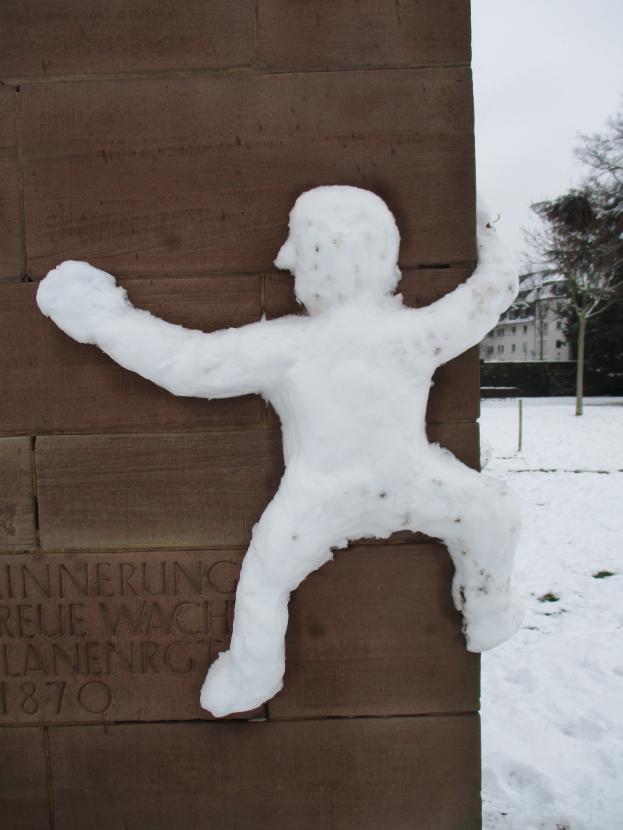 Eine Schneeskulptur eines Mannes an der Seite eines Geb├Ąudes, umgeben von B├Ąumen, Geb├Ąuden und einem klaren Himmel, mit Schnee drum herum.