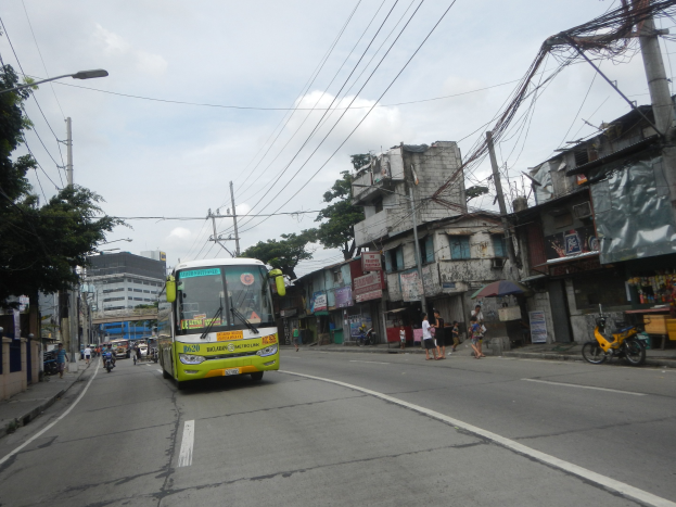 Grüner Bus fährt auf einer Straße mit hohen Gebäuden, Strommasten, Laternen, Bäumen, Fußgängern und Radfahrern unter einem bewölkten Himmel.