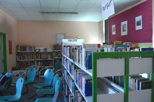 Ein Bibliotheksinneres mit Bücherregalen, Stühlen, Taschen auf dem Boden, gerahmten Kunstwerken an den Wänden, einer Anschlagtafel, einer Tür und Deckenleuchten.