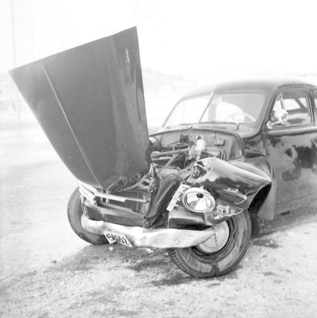 Schwarzes und weißes Foto eines schwerbeschädigten Autos am Straßenrand mit eingedellter Motorhaube, zersplitterten Fenstern und offener Fahrertür, die den Motor zeigt.