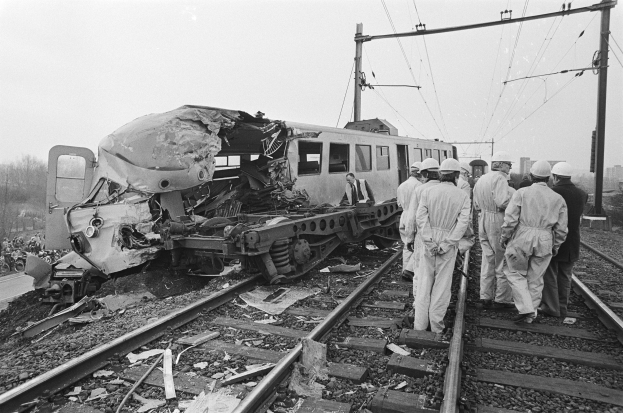 Schwarz-weiß-Bild einer Zugkollision auf den Gleisen mit Einsatzkräften in Helmen und einem Hintergrund aus Strommasten, Bäumen, Gebäuden und einem klaren Himmel.