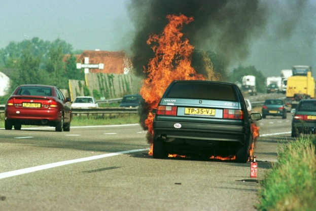 Ein Auto, das in Flammen steht, mit anderen Fahrzeugen in der Nähe, Bäumen und Gebäuden im Hintergrund bei klarem blauem Himmel, Gras auf der rechten Seite und einem Feuerlöscher.