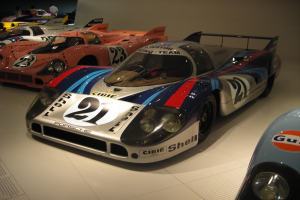 Ein Porsche 917K Le Mans-Rennwagen in einem Museum ausgestellt, umgeben von anderen Sportwagen, mit Menschen drumherum und einer Wand im Hintergrund, und ein Blatt Papier mit Text links daneben.