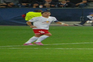 Ein Mann in weissem Hemd und roter Hose, der auf einem Fussballfeld Fussball spielt, umgeben von einer Gruppe von Menschen im Hintergrund.