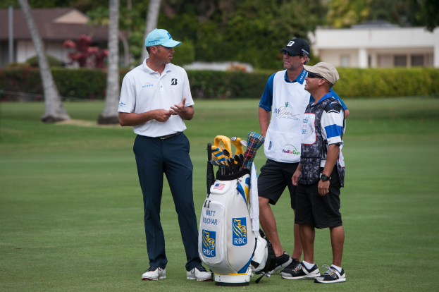 Eine Gruppe von Männern steht auf einem grünen Feld mit einem Golfbag im Vordergrund, Bäumen, Pflanzen und Gebäuden im Hintergrund, was auf ein Golfturnier hindeutet.