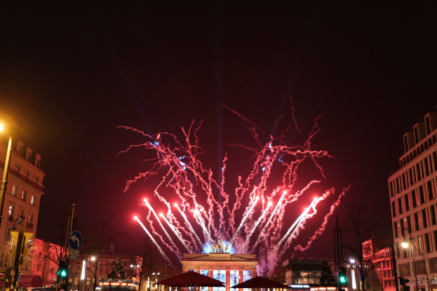 Eine belebte Stadtstraße an Silvester in Berlin, voller Menschen, Fahrzeuge und festlicher Beleuchtung von Gebäuden und Feuerwerk am Himmel.