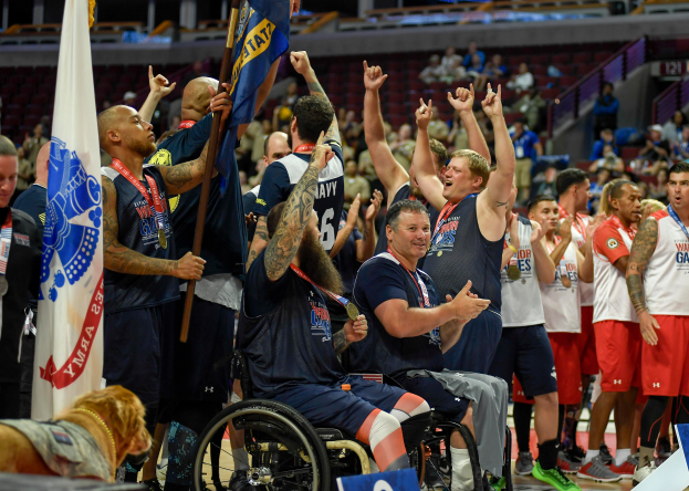 Männer im Rollstuhl auf einem Basketballfeld, einige halten Fahnen, feiernd mit einem Hund unten und Zuschauern im Hintergrund.