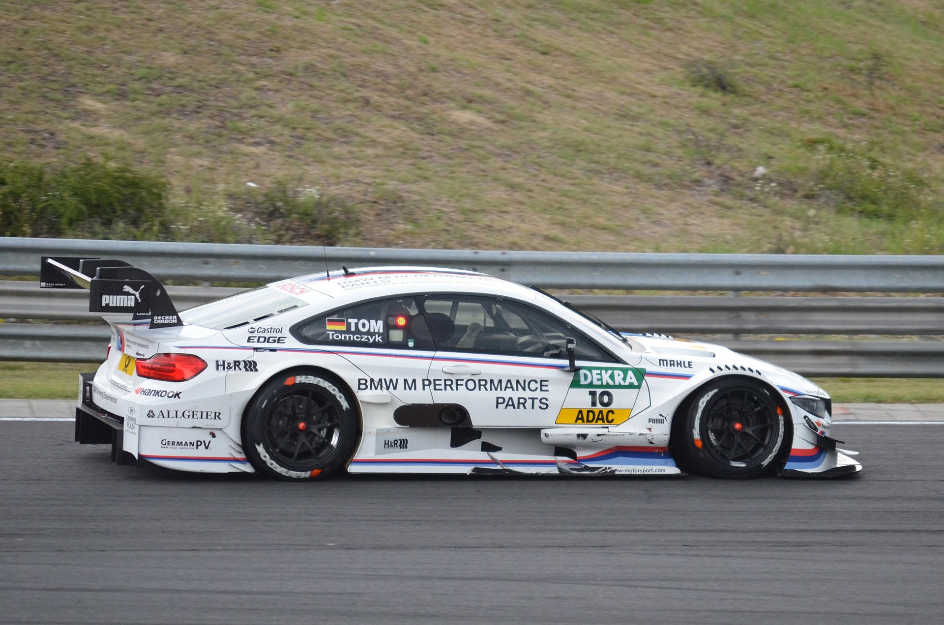 BMW M4 DTM Rennauto auf einer Strecke mit Text auf der Karosserie, umgeben von grünem Gras und einer Begrenzung im Hintergrund.