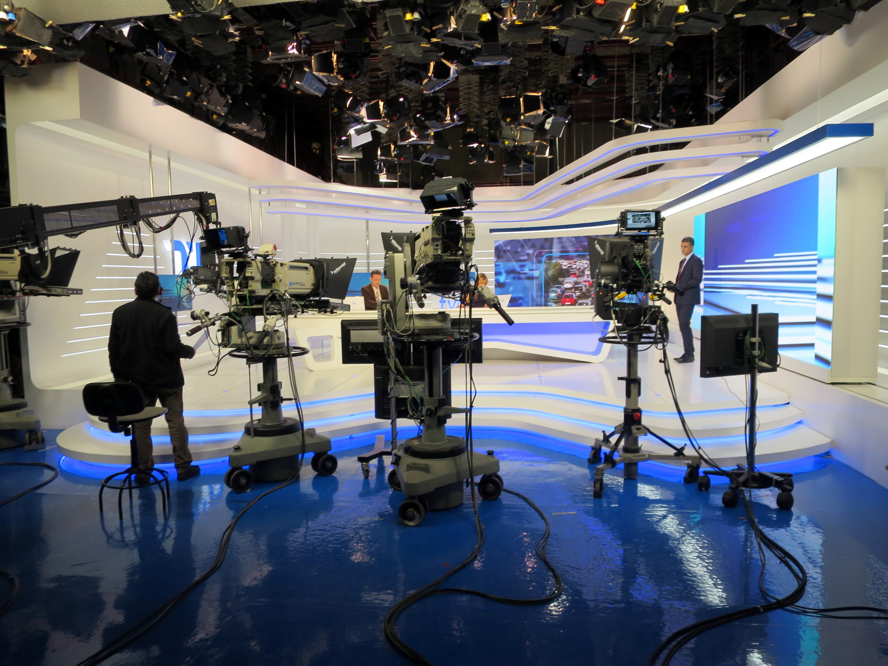 Ein TV-Studio mit mehreren Kameras auf dem Boden, einer sitzenden Person im Vordergrund und einigen stehenden Personen im Hintergrund, mit Deckenleuchten für eine Live-Übertragung.