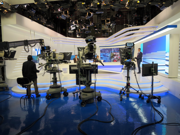 Ein TV-Studio mit mehreren Kameras auf dem Boden, einer sitzenden Person im Vordergrund und einigen stehenden Personen im Hintergrund, mit Deckenleuchten für eine Live-Übertragung.