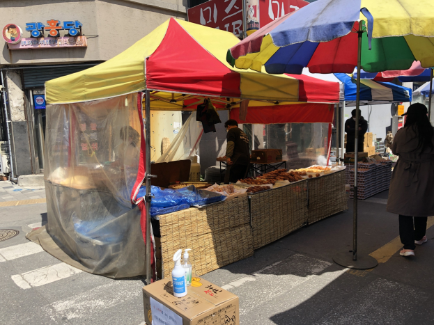 Ein Straßenhändler in Seoul, Südkorea, serviert Essen unter Zelten entlang einer belebten Straße, mit einer Kartonschachtel mit Flaschen auf der Straße und Gebäuden im Hintergrund unter einem klaren Himmel.